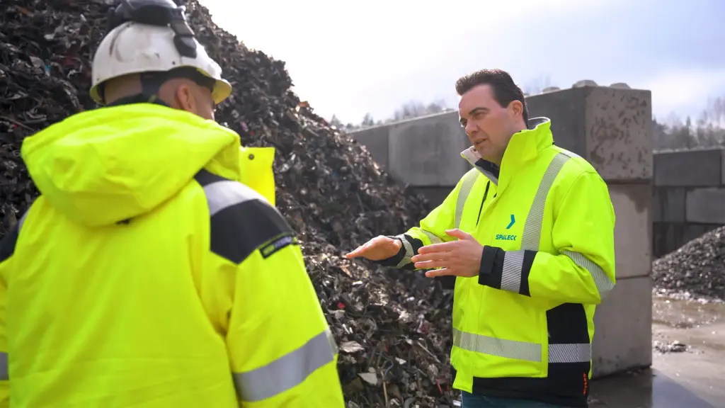 Two men in yellow jackets are talking in front of a pile of scrap metal.
