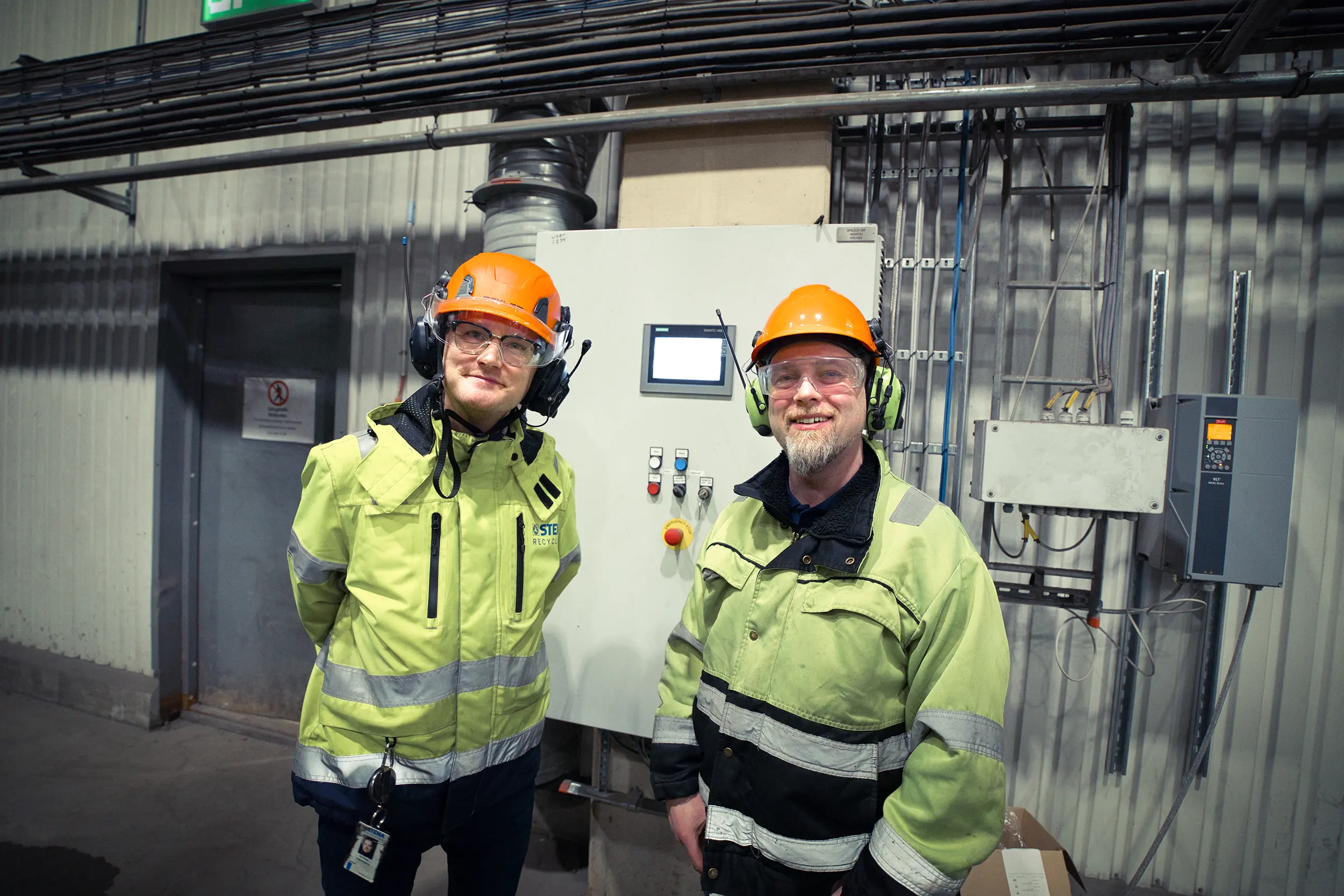 Two workers in protective gear standing in front of a control unit at a metal recycling facility.