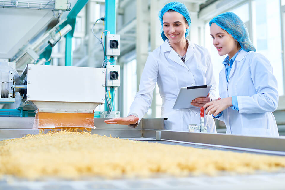 Two women in lab coats are inspecting a production line carrying bulk material.
